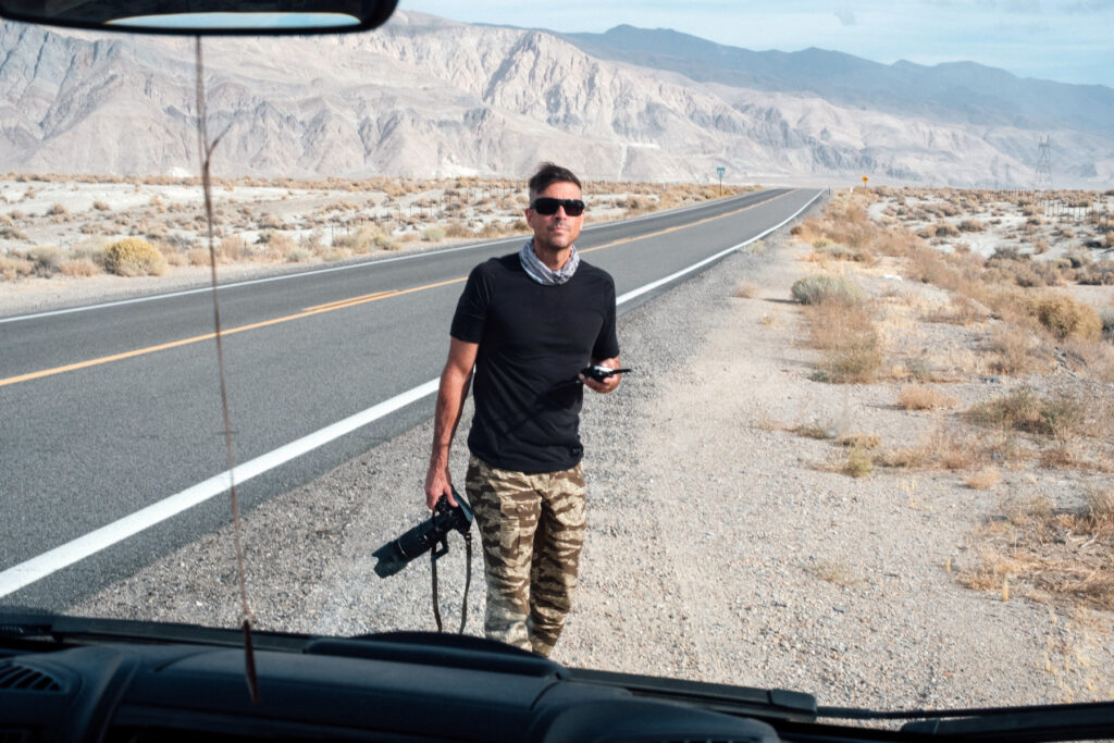 Photographer Dan Milnor stands on the side of a road in the desert with a large camera in hand.