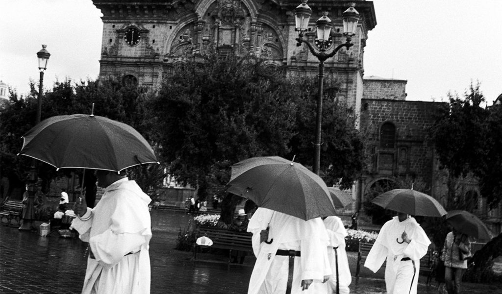 Four people wearing white robes holding umbrellas in front of a monument