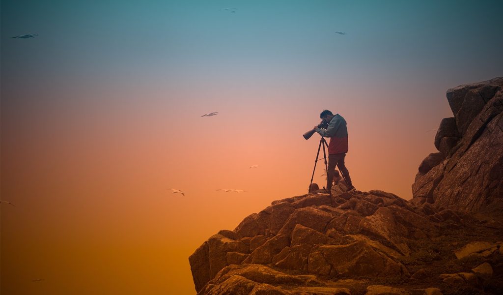 Photographer standing at the top of a rocky mountain taking a photo with a camera equipped with a long lens