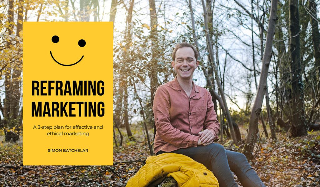 Simon Batchelar sitting outside on a log with a yellow jacket that matches the cover of their book, Reframing Marketing.