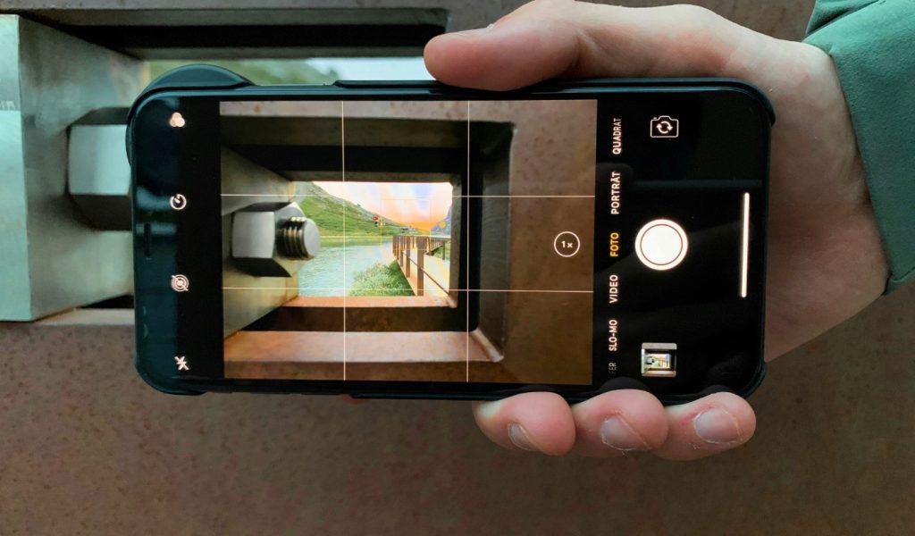 A hand holding a smartphone taking a photo of green hills and water through a gap in a wall, using on-screen grid lines to aid composition. 