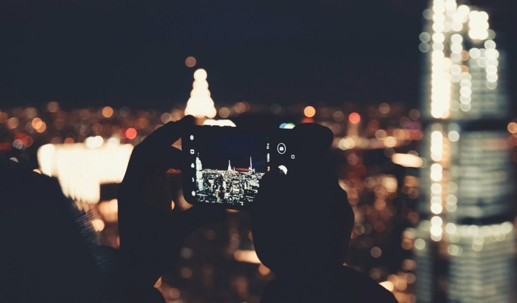 Person holding a smartphone at night, capturing a city skyline with illuminated buildings and blurred lights.